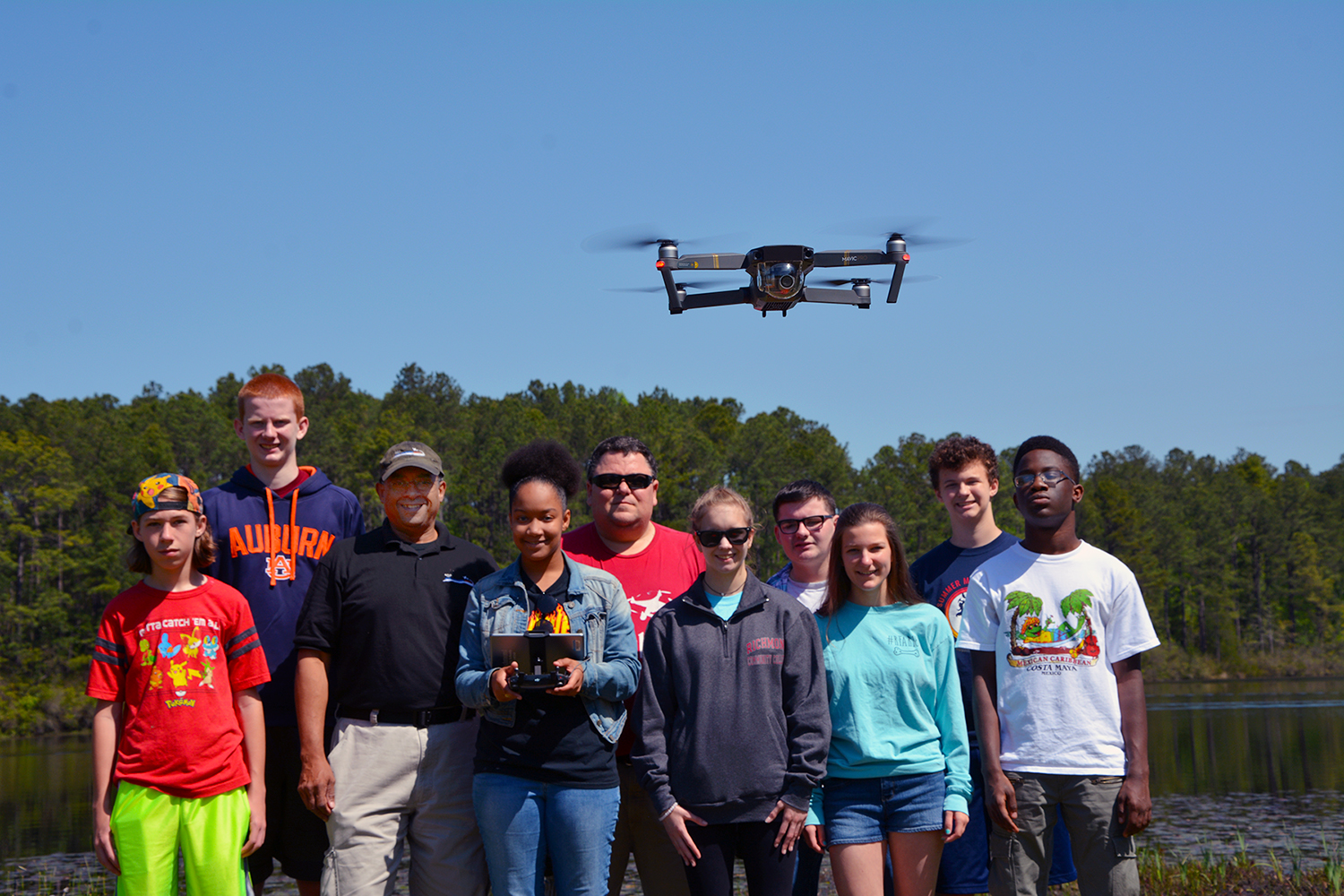 The Drone Academy students and instructors stand by the lake with a drone hovering in front of them.