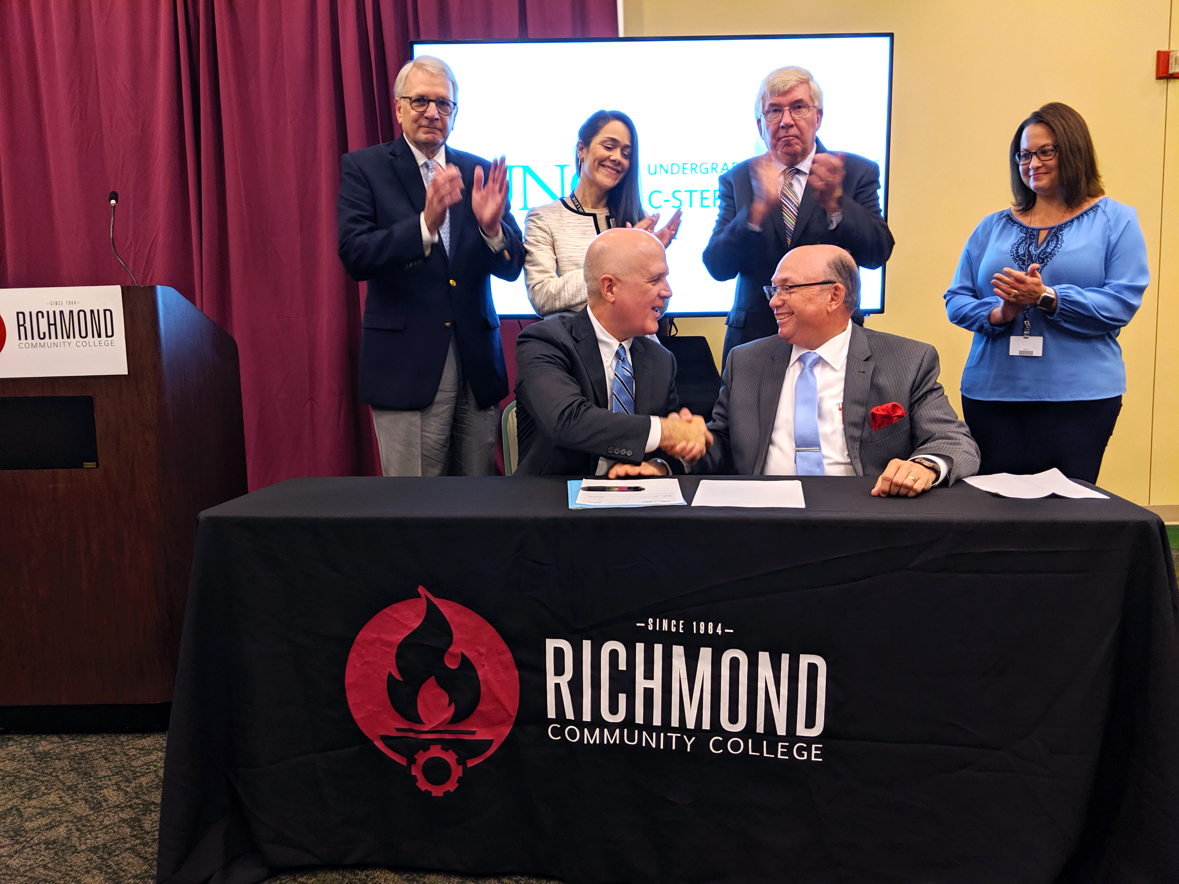 RichmondCC and UNC leaders sit at a table and sign a document while others look on.