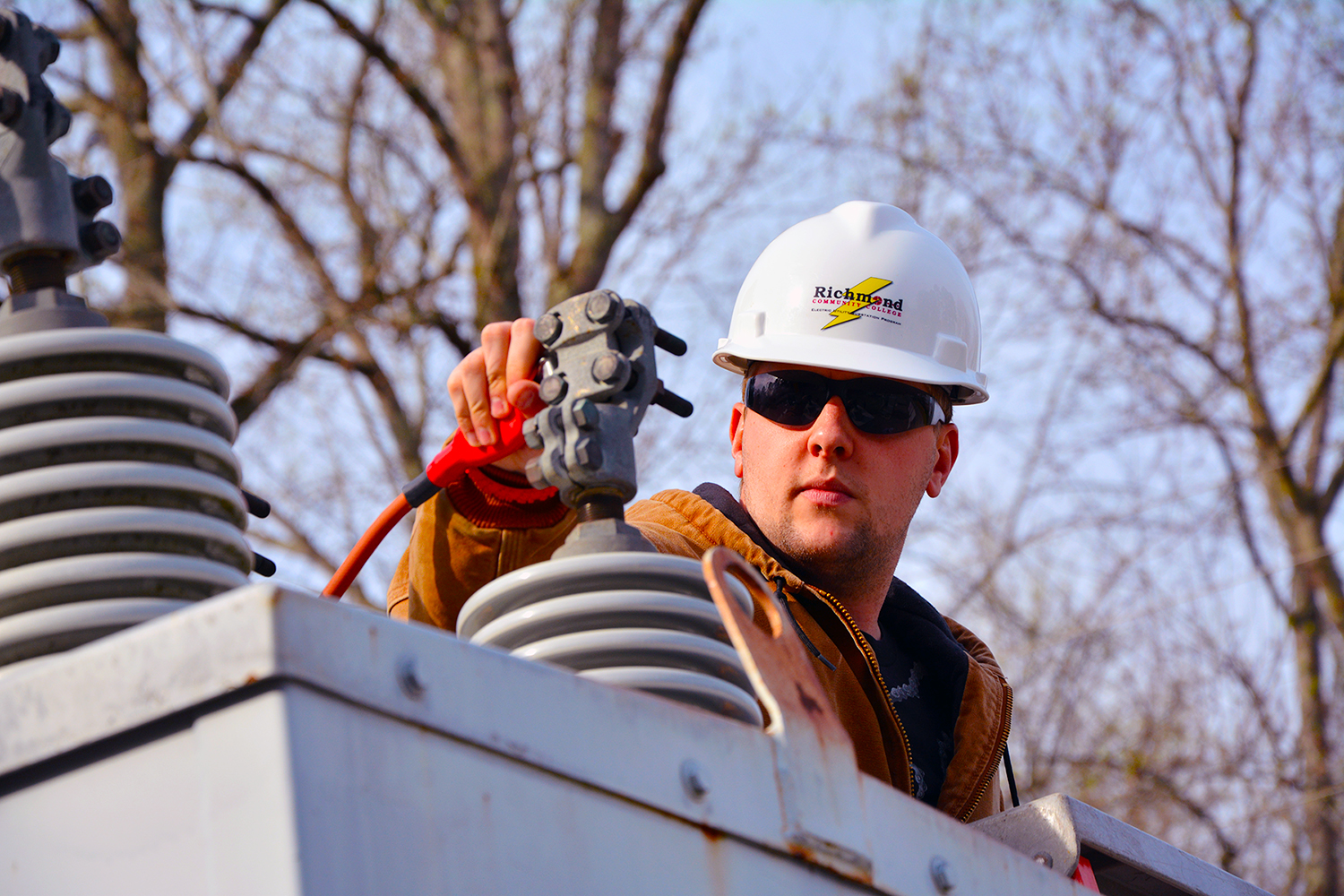A student wearing a hard hat works on a transformer in the substation.