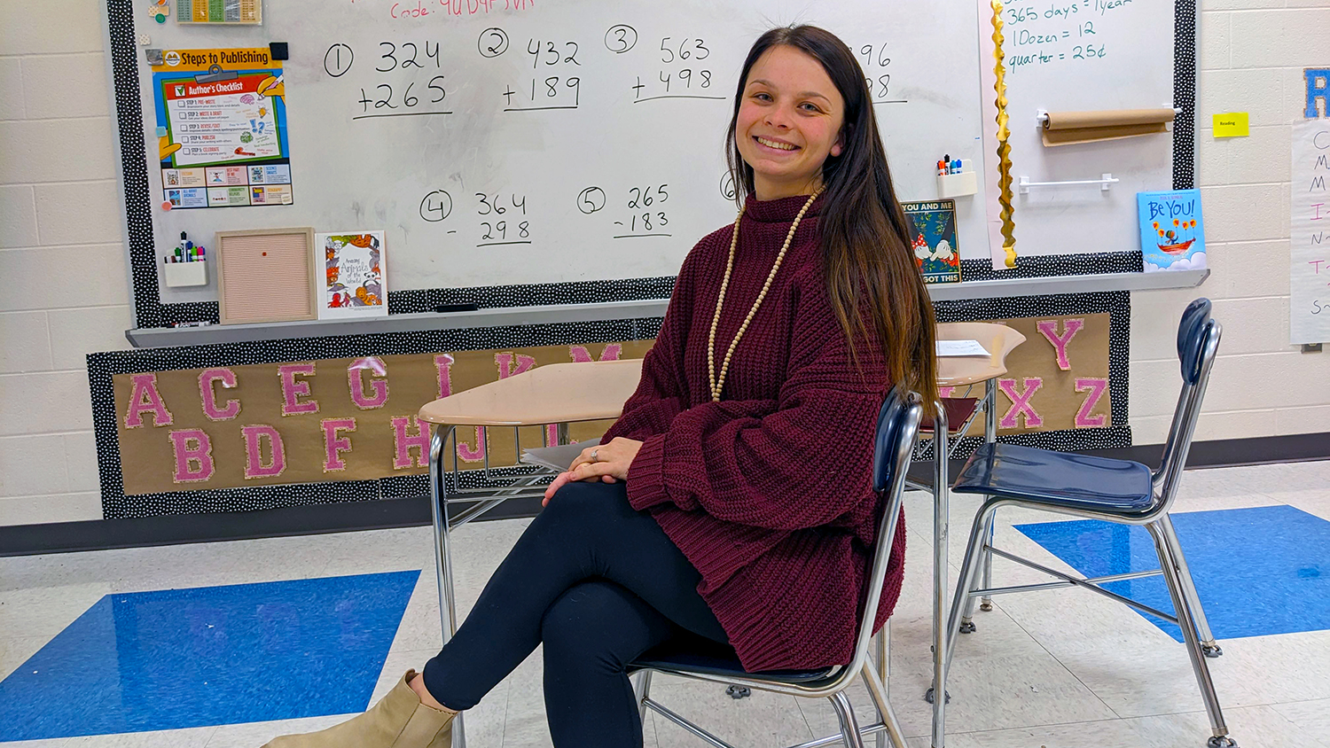 A student sits in a chair smiling. Behind her is a white board in a classroom.