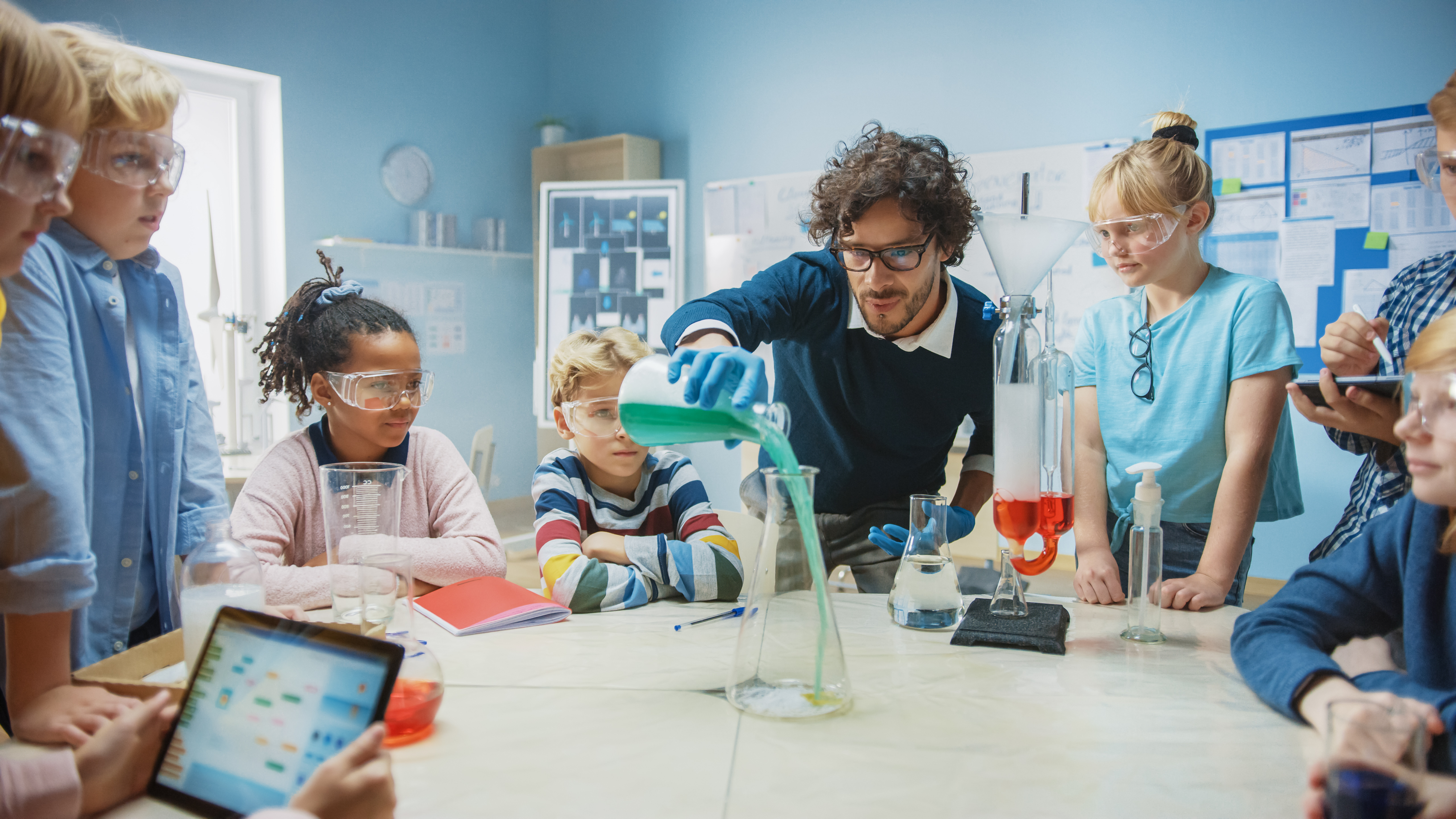 Image of a teacher pouring green liquid into a container while students watch and take notes.