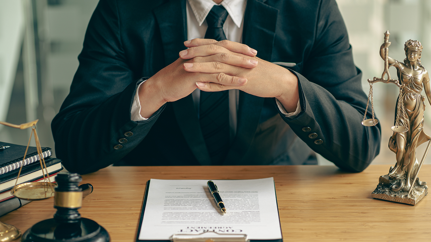 Shoulders and hands of a man in a suit sitting at a desk, an important document and pen on the table before him.