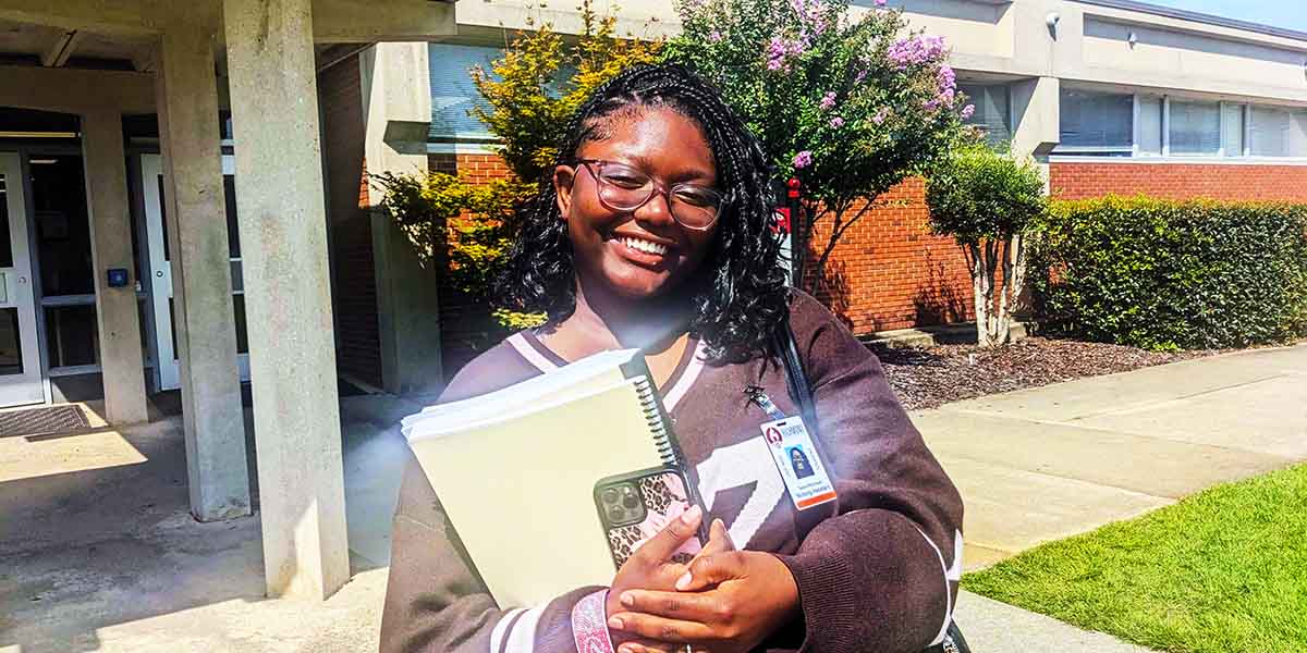 Student holding books and a cell phone and smiling for the camera.
