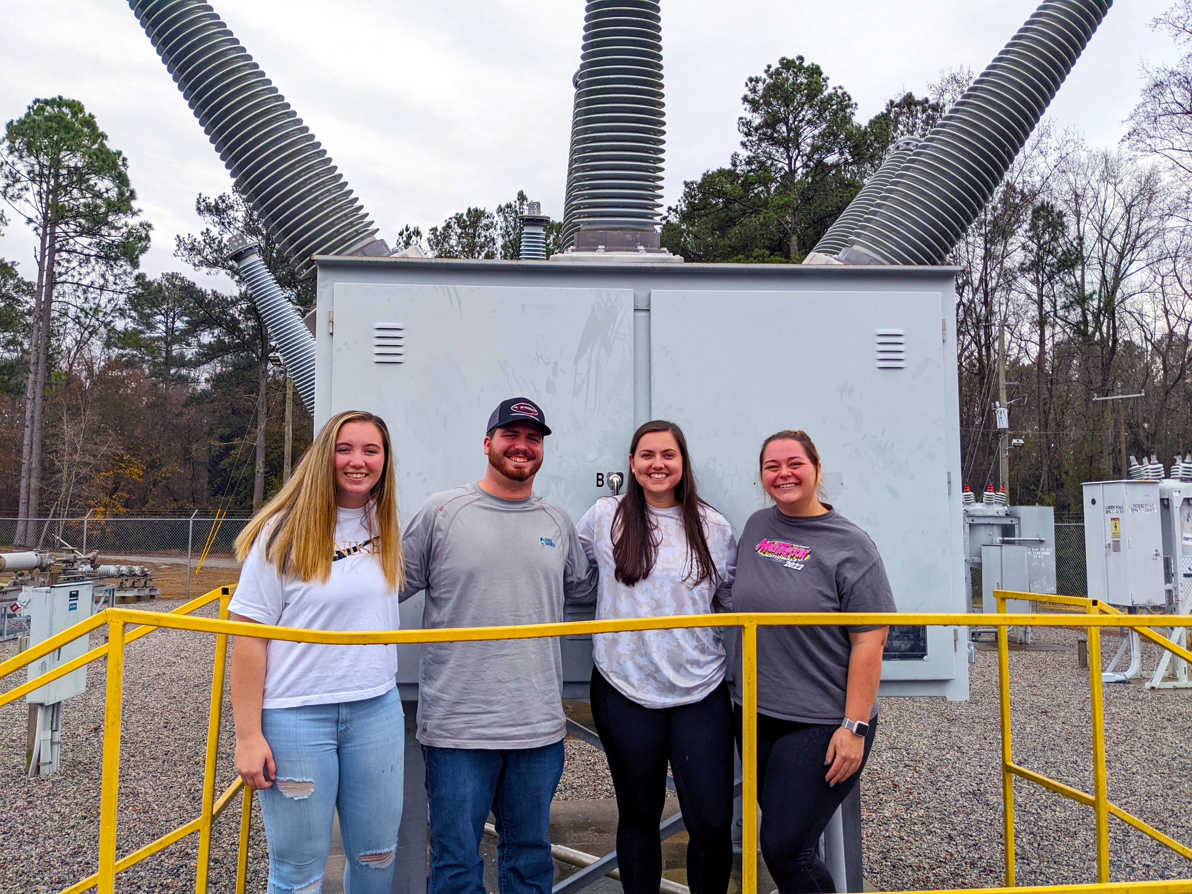 Four students stand in the campus substation lot with a transformer in the background.