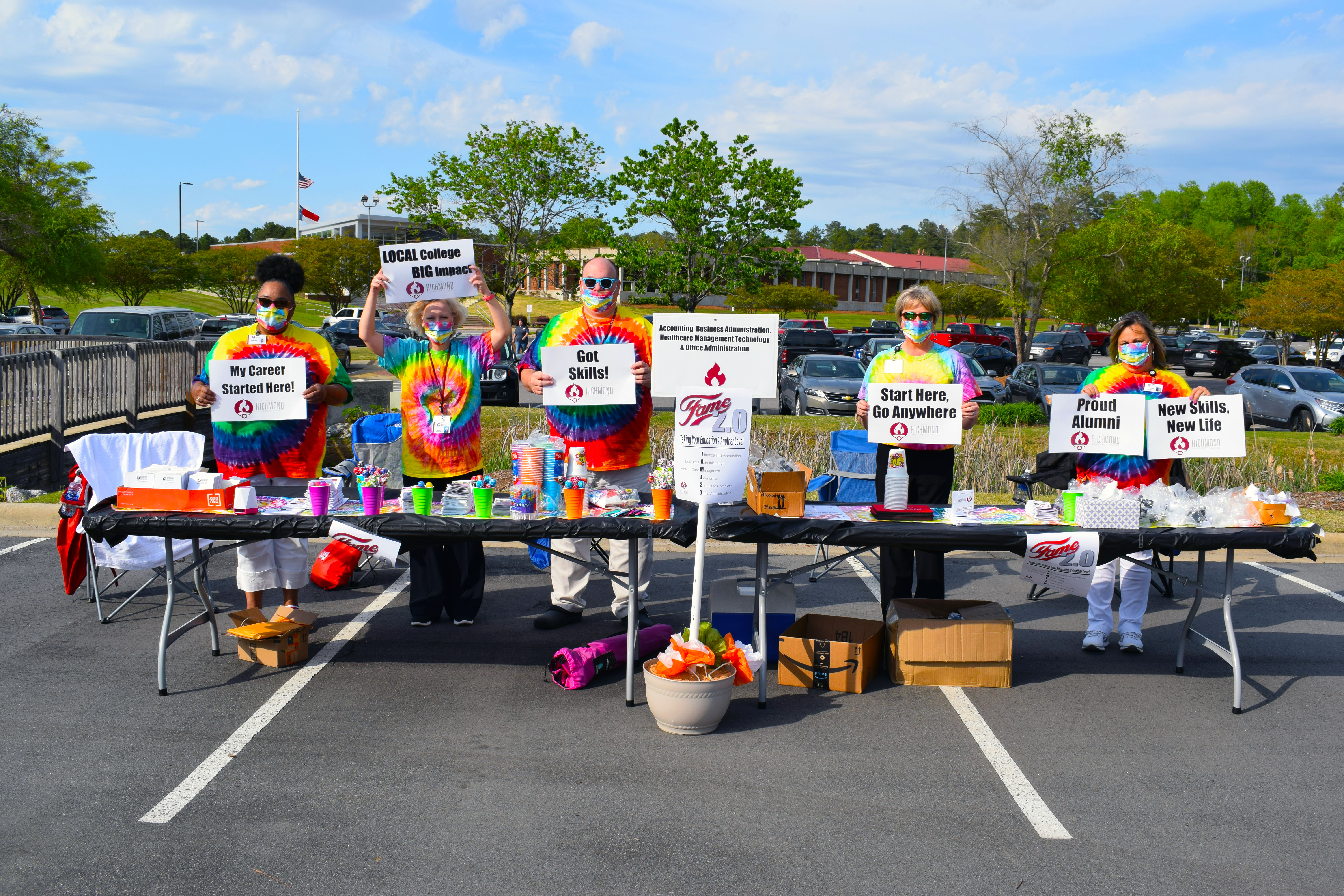 RichmondCC instructors in tye-dye shirts and masks stand at a table holding various signs about choosing RichmondCC.