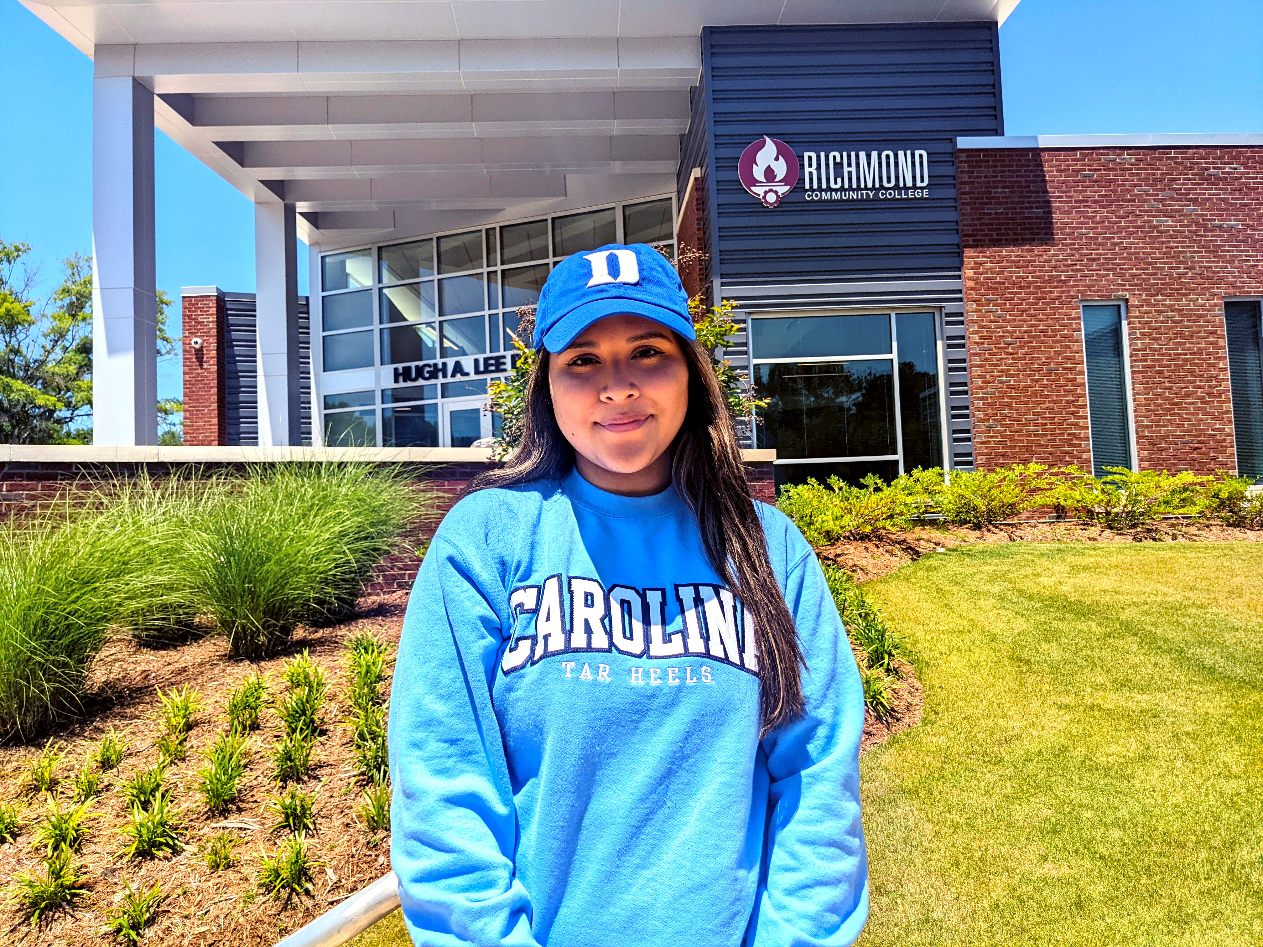 Carolina Mendez wears a Duke hat and a Carolina sweatshirt in front of the Lee Building with RCC logo in background.